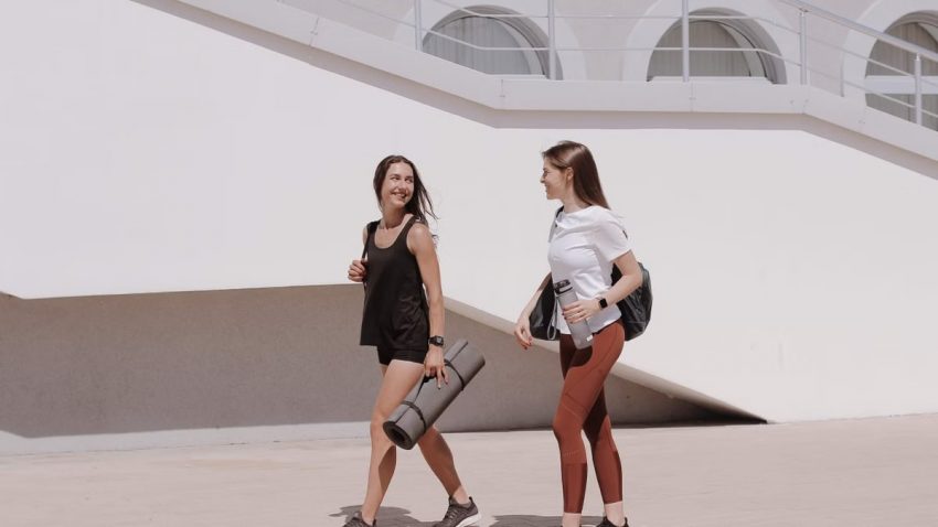 Two women in active wear walking outdoors while smiling and carrying yoga mats.